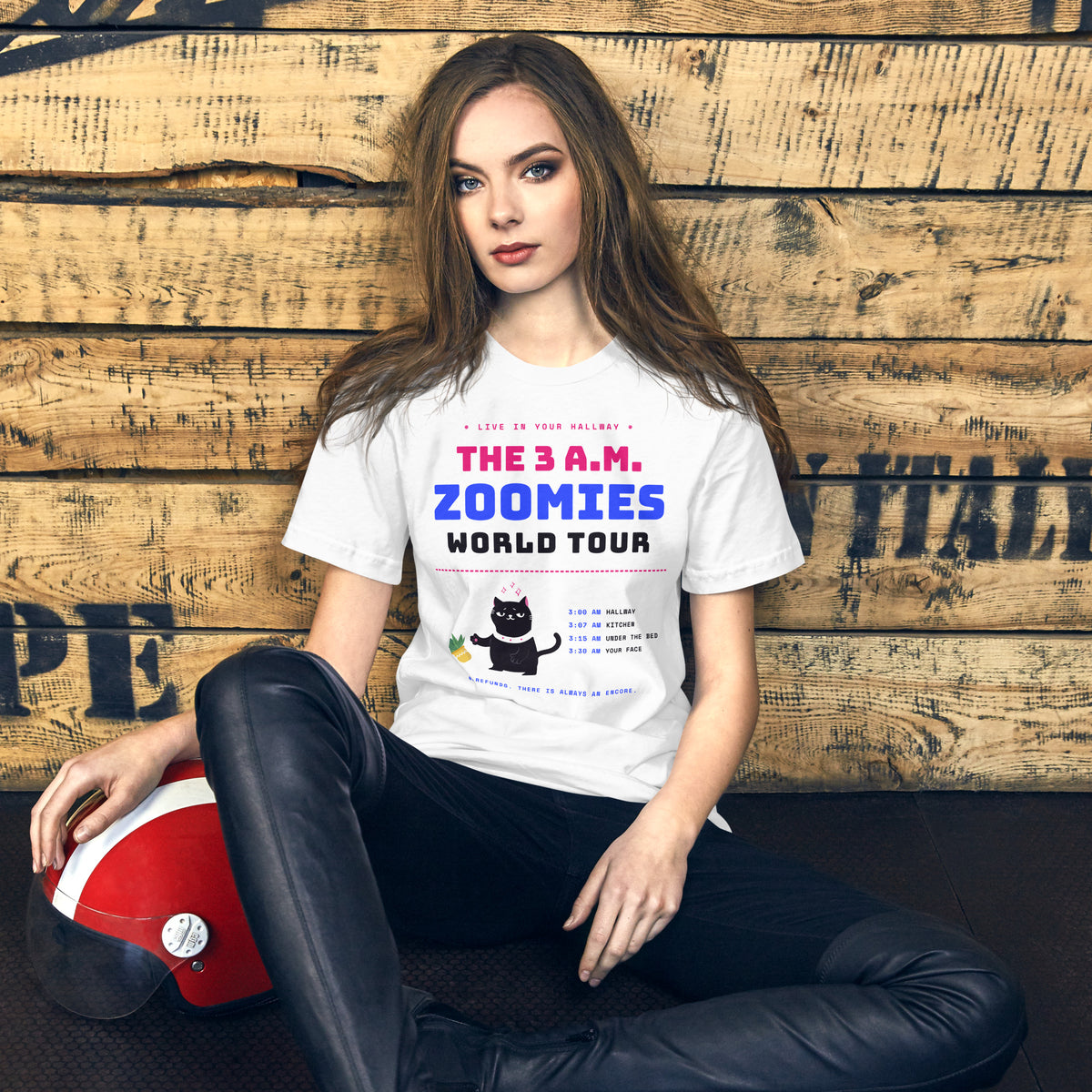 Woman wearing a white unisex t-shirt with a black cat knocking over a plant and the words “The 3 A.M. Zoomies World Tour” with a list of tour stops
